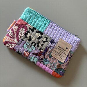 Könnte beinhalten: Eine gesteppte Reißverschlusstasche mit Patchwork-Design, das verschiedene florale und geometrische Muster in den Farben Lila, Rosa, Türkis und Grau zeigt. Die Tasche hat ein kleines beigefarbenes Etikett mit dem Text "little reminder YOU ARE AMAZING YOU CAN DO ANYTHING KISS UNCONTROLLABLY FALL IN LOVE DREAM FEARLESS BE YOURSELF, ALWAYS!"