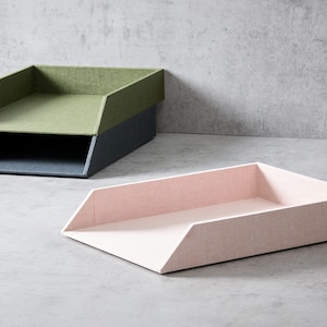 May include: Three modern desk organisers in shades of pink, green, and blue. The organisers have a rectangular shape with angled sides, and are made of a textured fabric material. The organisers are stacked on top of each other.