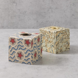 May include: Two square tissue boxes with decorative paper coverings. One box has a blue and red pattern with stars and waves. The other box has a marbled pattern in shades of green, brown, and white.