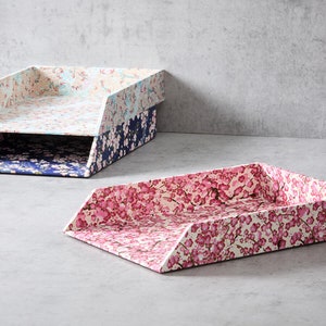 May include: Two decorative paper trays with a floral pattern. The top tray is light blue with a white floral pattern. The bottom tray is dark blue with a white floral pattern. The tray in the foreground is pink with a white floral pattern.