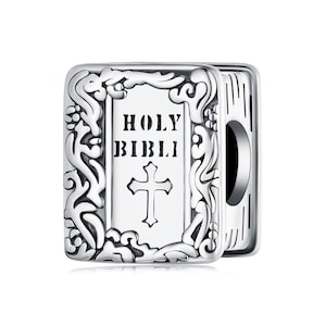 Puede incluir: Un charm plateado con forma de libro abierto. La portada dice "HOLY BIBLI" con una cruz debajo. El charm tiene adornos de volutas decorativas y está diseñado para usarse en una pulsera o collar.
