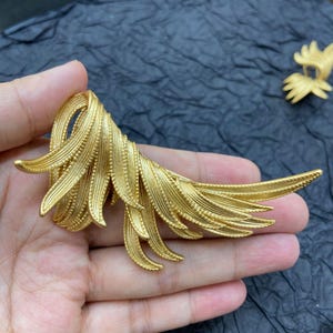 May include: A gold-colored, ornate brooch in the shape of a stylized feather or wing. The brooch has a detailed, textured surface with multiple layers and curves. The design is symmetrical and elegant, with a polished finish.