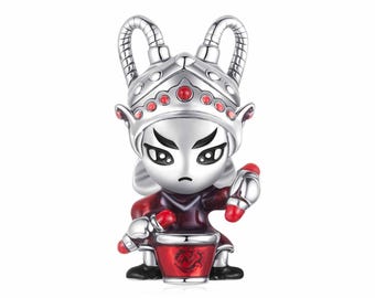 May include: A silver-colored charm depicting a stylized warrior figure. The charm has a red and silver helmet, red accents, and is holding a red drum. The figure is in a seated position.