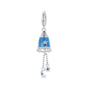 May include: A silver charm in the shape of a blue rocket with a white star and a dangling moon and star charm. The rocket is decorated with clear crystals.
