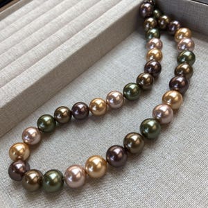 Op de afbeelding: Een veelkleurige parelketting met tinten goud, bruin, groen en roze. De ronde parels zijn geregen, waardoor een klassiek sieraad ontstaat. De ketting wordt gepresenteerd op een neutraal getinte ondergrond. De parels zijn ongeveer 0,8 cm in diameter.