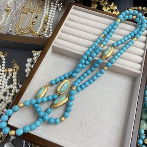 Może przedstawiać: Turkusowy naszyjnik z koralików ze złotymi akcentami. Naszyjnik składa się z trzech sznurów okrągłych turkusowych koralików, przeplatanych złotymi koralikami oraz owalnymi turkusowo-złotymi zawieszkami. W tle widoczna jest inna biżuteria.