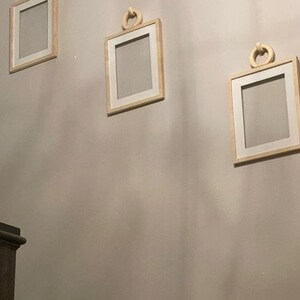 Puede incluir: Tres marcos de fotos de madera con paspartús blancos y asas de cuerda decorativas cuelgan de una pared gris claro. Los marcos están dispuestos en forma escalonada.