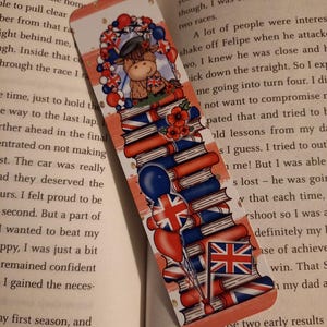 May include: A rectangular bookmark with a rounded top, decorated with a cartoon Highland cow in a beret, balloons, and a wreath. The design includes stacks of books, the Union Jack flag, and red poppies. The bookmark is designed to be placed in a book.
