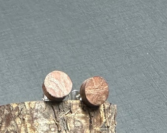 Könnte beinhalten: Ein Paar kreisförmige Ohrstecker mit einer natürlichen braunen Holzmaserung. Die Ohrringe haben eine flache, runde Vorderseite mit sichtbarer Holzstruktur und sind an silberfarbenen Metallstiften befestigt, präsentiert auf einem Stück rauer Rinde.
