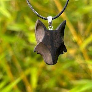 Può includere: Un ciondolo in legno marrone scuro a forma di testa di volpe, sospeso a un cordino nero. Il ciondolo è scolpito con dettagli e pende da un'anello color argento. Lo sfondo è una vista sfocata di fogliame verde.