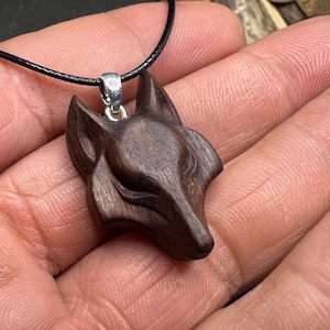 May include: A dark brown wooden pendant carved into the shape of a fox head. The pendant features a smooth, polished finish with visible wood grain and is attached to a simple black cord by a small, silver-coloured metal bail at the top.
