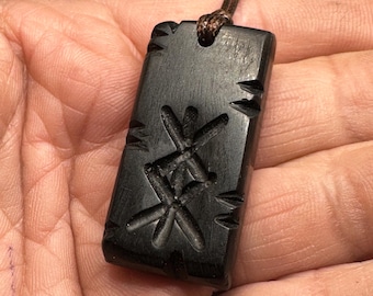 May include: A rectangular black pendant with a smooth finish, featuring two carved intersecting linear symbols on the front surface and small notches along the edges. The pendant is suspended from a thin, dark brown cord.