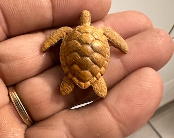 May include: A small, intricately carved wooden turtle figurine. The turtle is a warm, light brown color with detailed shell patterns and outstretched flippers. The figurine is a miniature representation of a sea turtle.