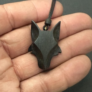Può includere: Una collana con pendente a forma di volpe grigio scuro. Il pendente è scolpito a forma di testa di volpe con orecchie a punta. La collana ha un cordino nero. Il pendente è tenuto in mano.