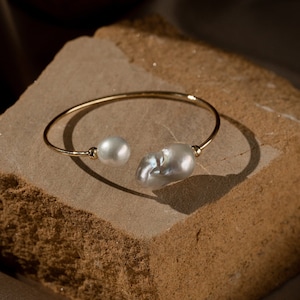May include: A gold bangle bracelet with two irregular shaped pearls. The bracelet is on a brown textured surface.