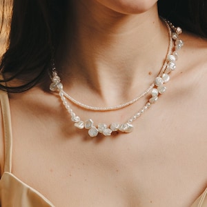 May include: A woman with long dark hair wearing a beige tank top and a layered pearl necklace.