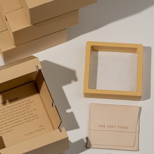 May include: A set of packaging for jewelry. The packaging includes a brown cardboard box with the text "THE VERY PEARL" embossed on the top, a smaller brown box with a white interior, a beige pouch with the text "THE VERY PEARL" printed on the front, and a small beige cleaning cloth with the text "THE VERY PEARL" printed on it.
