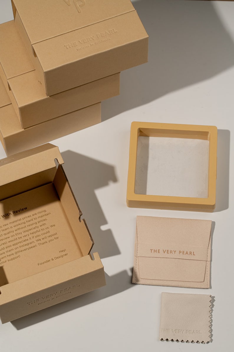 May include: A set of packaging for jewelry. The packaging includes a brown cardboard box with the text "THE VERY PEARL" embossed on the top, a smaller brown box with a white interior, a beige pouch with the text "THE VERY PEARL" printed on the front, and a small beige cleaning cloth with the text "THE VERY PEARL" printed on it.