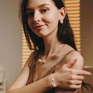 May include: A woman wearing a brown knitted tank top with a gold chain necklace and bracelet featuring pearl accents. The necklace and bracelet have three pearls each.