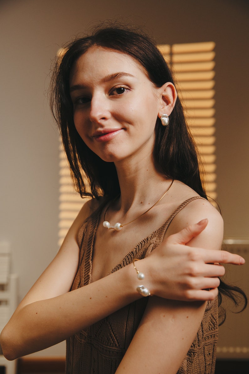 May include: A woman wearing a brown knitted tank top with a gold chain necklace and bracelet featuring pearl accents. The necklace and bracelet have three pearls each.