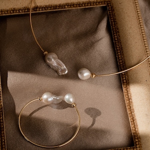 May include: Two gold necklaces with pearl accents. The necklaces are displayed on a brown surface inside a gold picture frame.