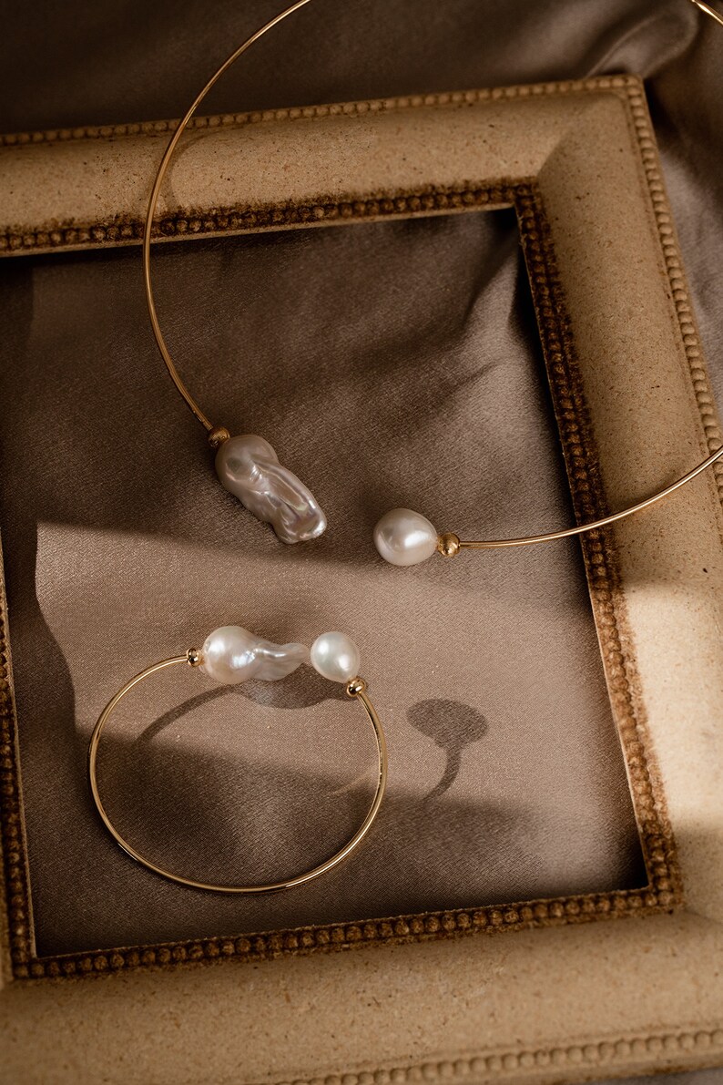 May include: Two gold necklaces with pearl accents. The necklaces are displayed on a brown surface inside a gold picture frame.