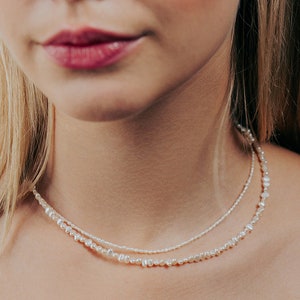 May include: A woman wearing a layered pearl necklace. The necklace is made of small, white pearls and is layered with a thinner chain of smaller pearls.