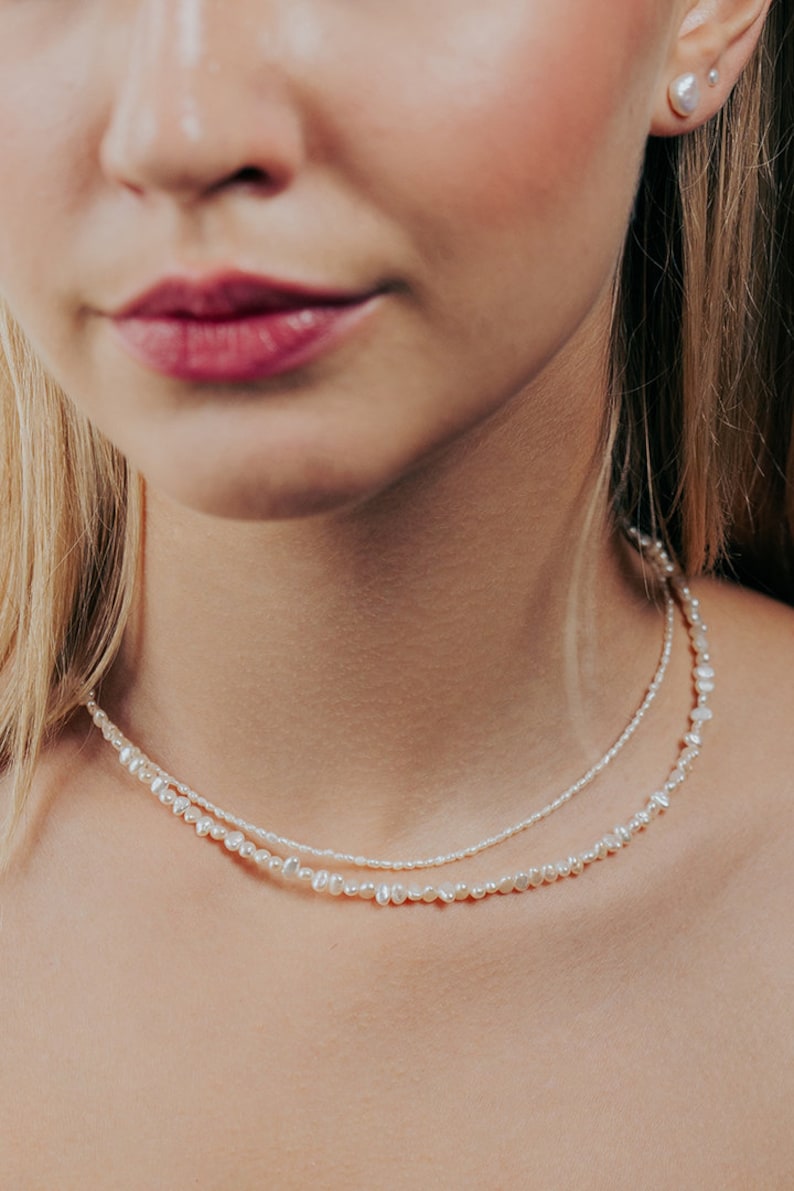 May include: A woman wearing a layered pearl necklace. The necklace is made of small, white pearls and is layered with a thinner chain of smaller pearls.