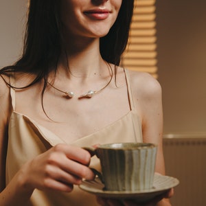 May include: A woman wearing a beige satin top with a gold and pearl necklace holds a green and white ceramic teacup and saucer.