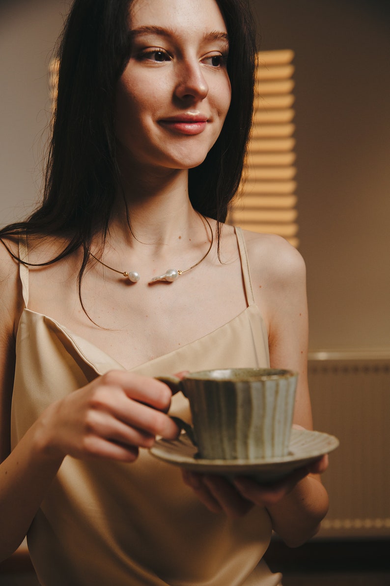 May include: A woman wearing a beige satin top with a gold and pearl necklace holds a green and white ceramic teacup and saucer.