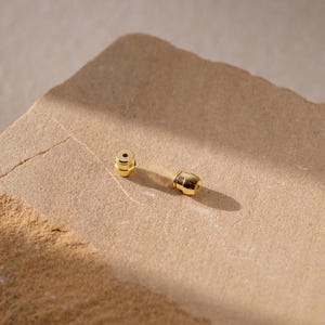May include: Two small, polished gold-tone earring backs with a cylindrical shape and a central hole for a post. These metallic jewelry components feature a smooth, reflective surface and are designed to secure earrings firmly in place.