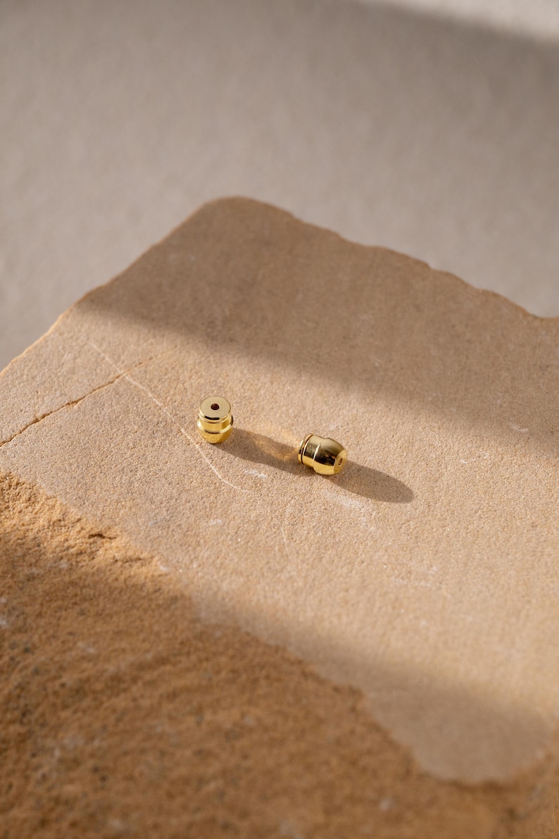 May include: Two small, polished gold-tone earring backs with a cylindrical shape and a central hole for a post. These metallic jewelry components feature a smooth, reflective surface and are designed to secure earrings firmly in place.