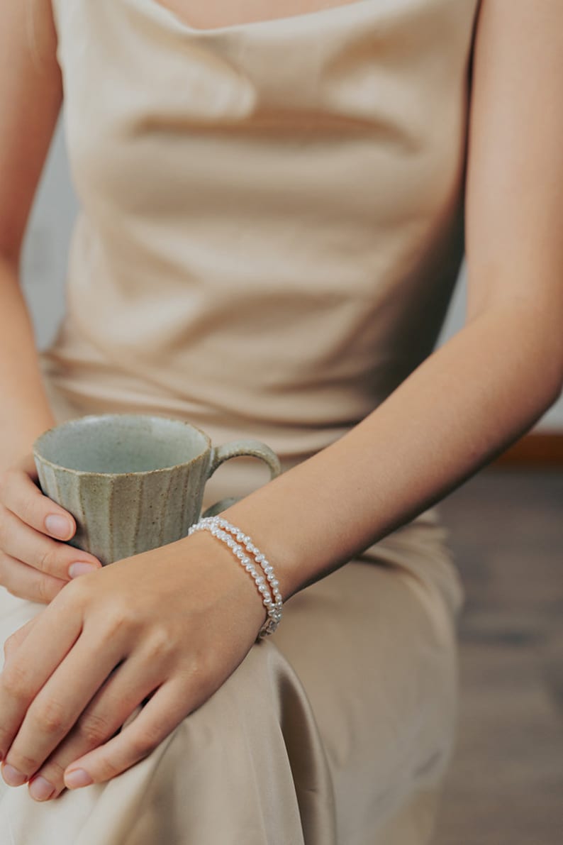 May include: A woman wearing a beige satin dress with a double strand pearl bracelet on her wrist.