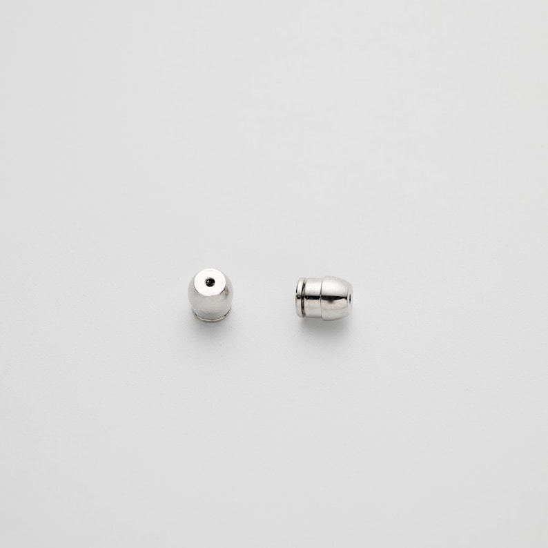May include: Two silver-toned metal earring backs with a bullet-shaped design. Each piece features a smooth, polished surface and a small central opening intended to secure an earring post, presented in both a front-facing and side-profile view.