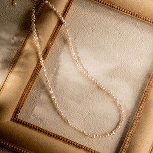 May include: A delicate pearl necklace with a gold clasp, displayed on a light brown picture frame.