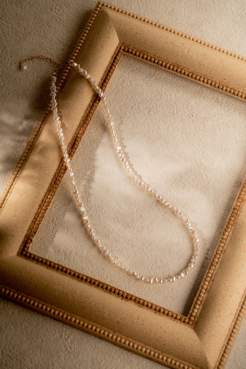 May include: A delicate pearl necklace with a gold clasp, displayed on a light brown picture frame.