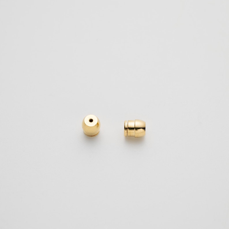 May include: Two small, cylindrical gold-toned earring backs. One is shown from the top, revealing a circular opening, while the other is positioned on its side, displaying a smooth, metallic surface with a subtle grooved detail near the base.