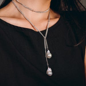 May include: A silver necklace with two strands, one a choker and the other a longer lariat style. The lariat features two large, irregularly shaped, white pearls. The necklace is worn against a black top.