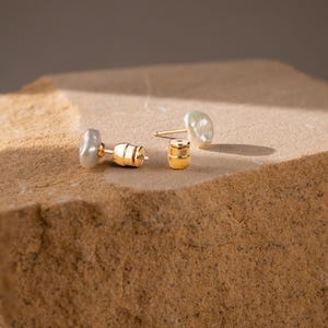 May include: A pair of stud earrings featuring irregular, off-white baroque pearls attached to gold-toned metal posts. The set includes two matching gold-toned metal earring backs, each with a cylindrical shape and a small central hole.