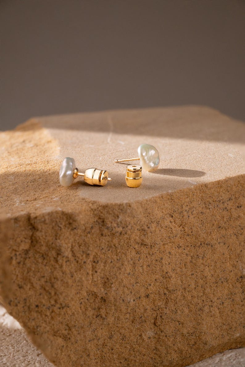 May include: A pair of stud earrings featuring irregular, off-white baroque pearls attached to gold-toned metal posts. The set includes two matching gold-toned metal earring backs, each with a cylindrical shape and a small central hole.