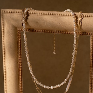May include: A delicate pearl necklace with a gold chain, both hanging in a wooden picture frame. The pearl necklace is made of small, irregular pearls, and the gold chain is made of linked oval shapes.