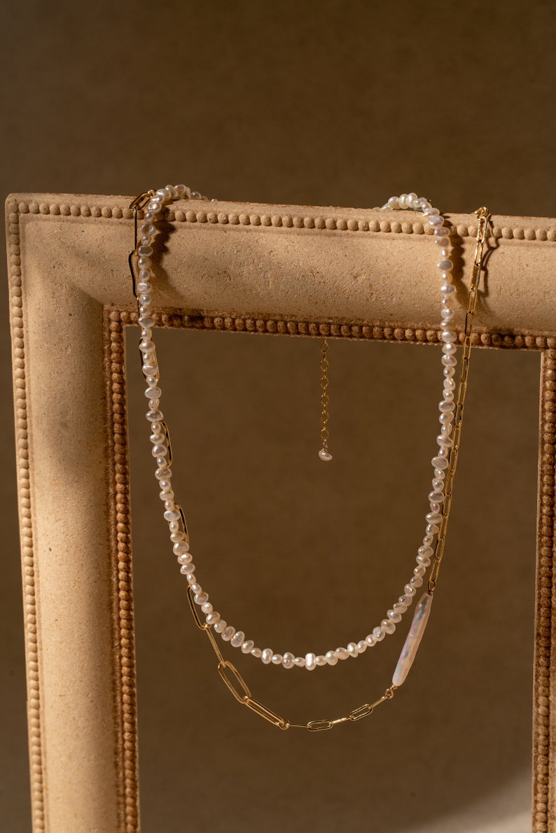 May include: A delicate pearl necklace with a gold chain, both hanging in a wooden picture frame. The pearl necklace is made of small, irregular pearls, and the gold chain is made of linked oval shapes.