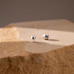May include: Two small, polished silver-toned earring backs with a cylindrical shape and a flat top. These metallic components feature a smooth, reflective surface and a central opening, designed to secure earrings firmly in place.