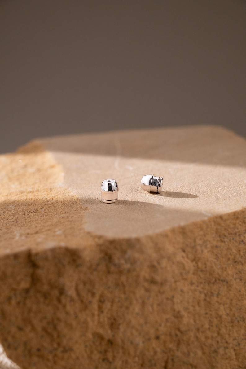 May include: Two small, polished silver-toned earring backs with a cylindrical shape and a flat top. These metallic components feature a smooth, reflective surface and a central opening, designed to secure earrings firmly in place.