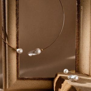 May include: Two gold necklaces with pearl accents. One necklace is displayed in a wooden frame, while the other is on a stone surface.