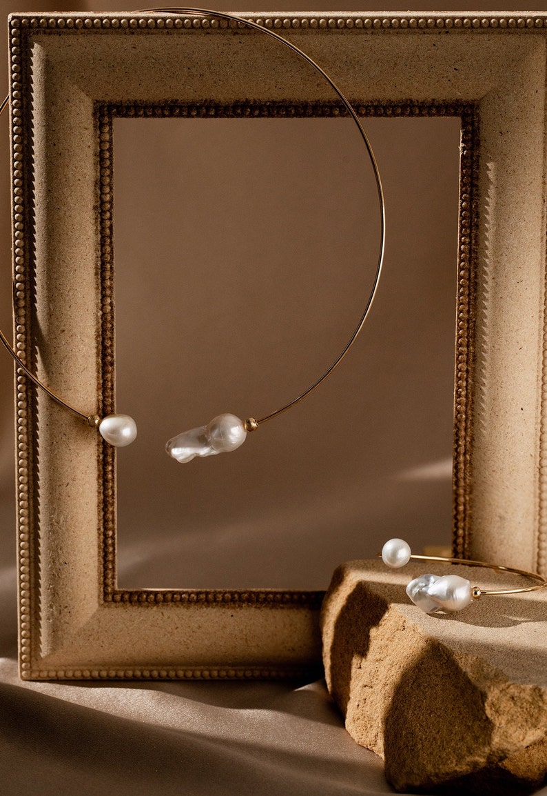 May include: Two gold necklaces with pearl accents. One necklace is displayed in a wooden frame, while the other is on a stone surface.