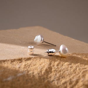 May include: A pair of white baroque pearl stud earrings with silver posts and matching silver earring backs. The pearls have an irregular, organic shape with a smooth, lustrous surface. The earrings are displayed as separate components.