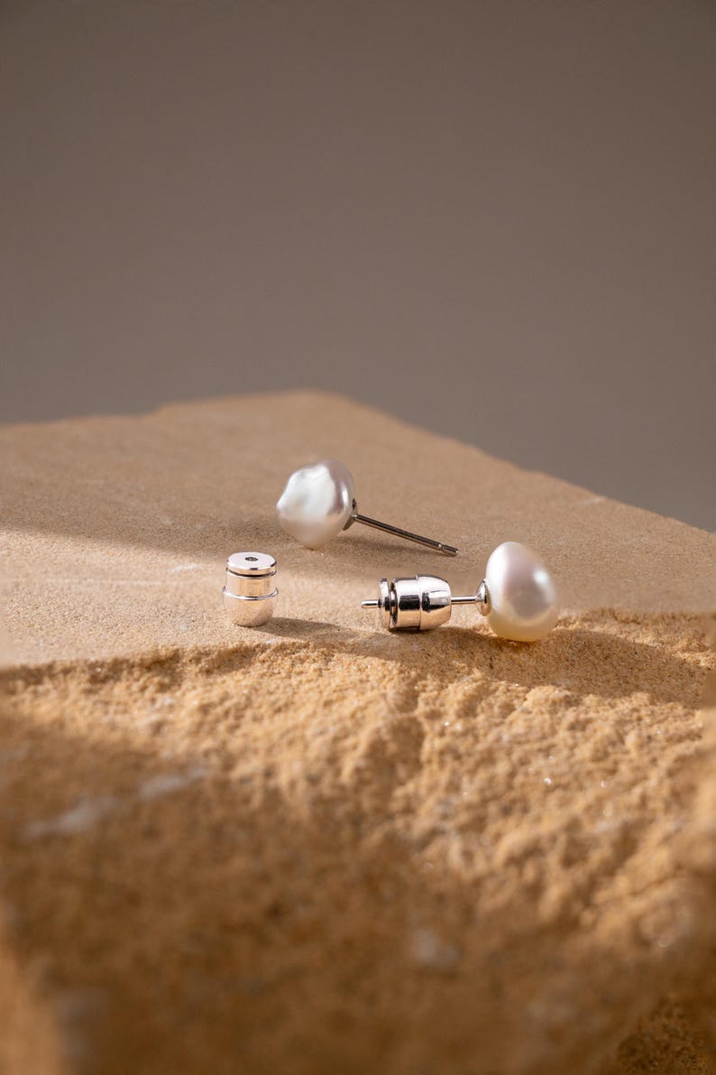 May include: A pair of white baroque pearl stud earrings with silver posts and matching silver earring backs. The pearls have an irregular, organic shape with a smooth, lustrous surface. The earrings are displayed as separate components.