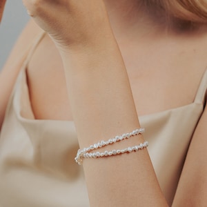 May include: A double-strand pearl bracelet with small, irregular pearls. The bracelet is made of white pearls and is worn on a person's wrist.