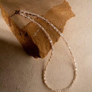 May include: A white pearl necklace with a gold clasp, laying on a brown rock.